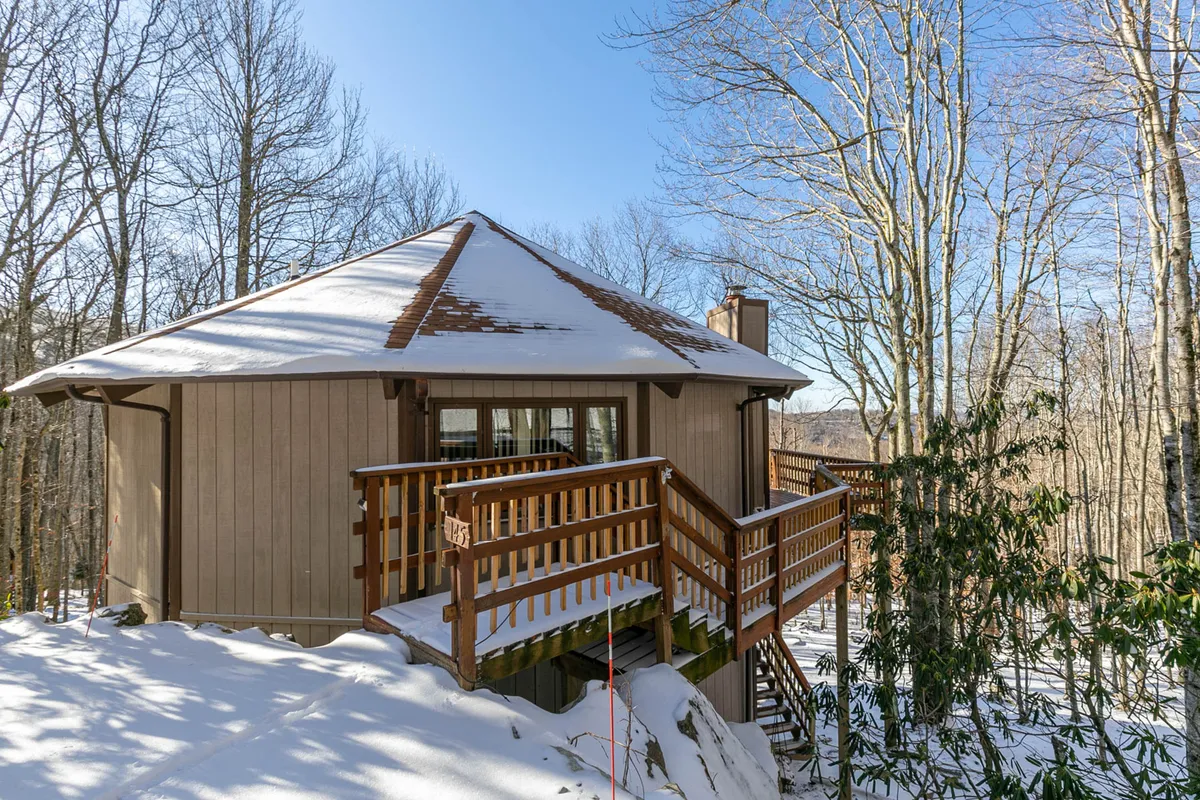 A peaceful winter scene showing a wooden deck overlooking snow-covered ground and surrounding trees, with the unique roundhouse roofline visible. The placement of the deck amid nature suggests a seamless blend between indoor comfort and the wild outdoors.