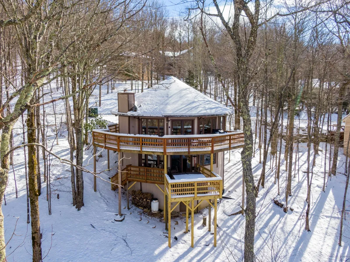 The hero image showcases the round, mountain cabin perched atop a snowy landscape, nestled among tall evergreens. Two spacious decks wrap around the home, inviting guests to enjoy panoramic, snow-dusted mountain views. The serene setting highlights the inviting warmth of the house against the crisp winter scene, promising cozy relaxation and outdoor adventure.