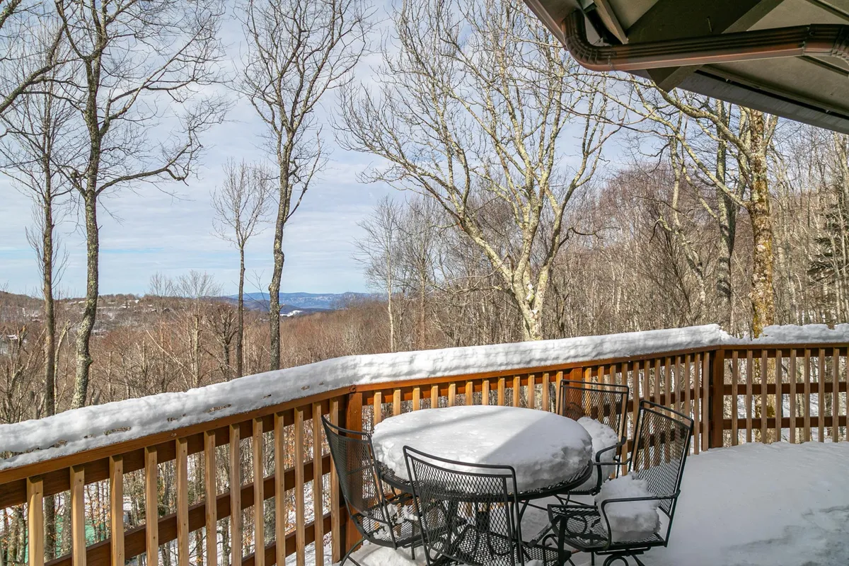 This exterior view features a wooden deck railing, a snow-covered table, bare trees, and distant mountains. It suggests gatherings outside, enjoying morning coffee or evening sunsets, and provides a strong sense of place with its mountain backdrop.