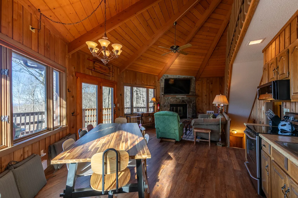 A panoramic view of the open-concept living and dining area inside the roundhouse, featuring a large wooden dining table set for eight, a striking stone fireplace as the focal point, and floor-to-ceiling windows framing vibrant sunset mountain vistas. The plush sectional and chandelier add a sense of warmth and elegance.