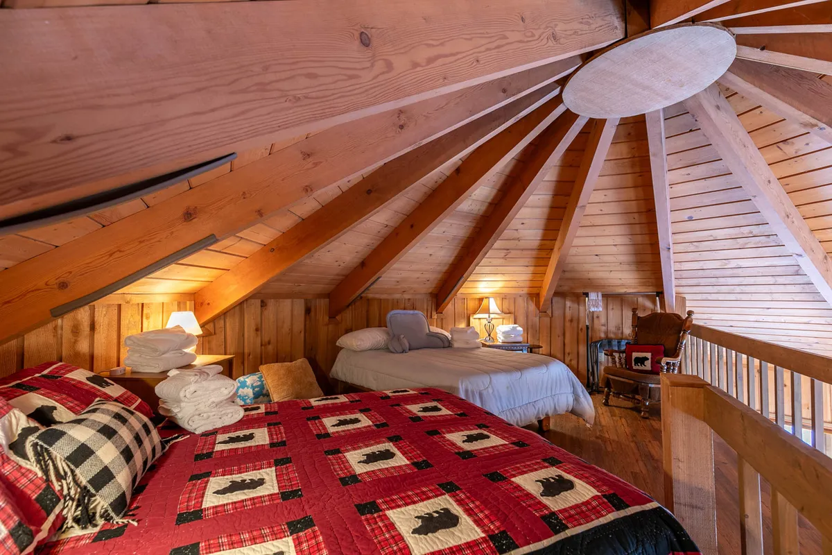 A cozy sleeping area featuring two beds—one with a red quilt and another with grey bedding—set beneath exposed wooden beams. There is a rocking chair in the corner and a circular window that lets in natural light, enhancing the airy feel of the loft. The space is intimate yet open, inviting both relaxation and playful moments.
