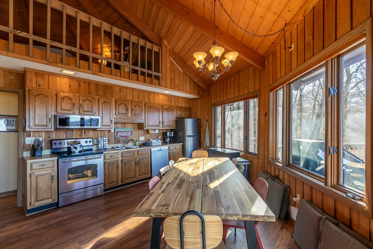 A bright interior shot focusing on the transition between the kitchen and dining areas. The fully equipped modern kitchen is visible, with stainless appliances, a wooden dining table, and large windows that draw the outdoors in. The sense of flow between kitchen, dining, and living spaces is apparent.