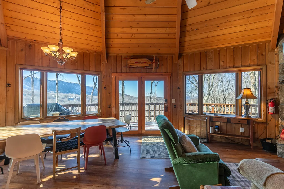 A spacious, inviting open living area with a large wooden dining table, a comfortable seating zone with plush sofas, and floor-to-ceiling windows that flood the space with natural light. The chandelier and warm decor evoke a sense of togetherness and celebration.