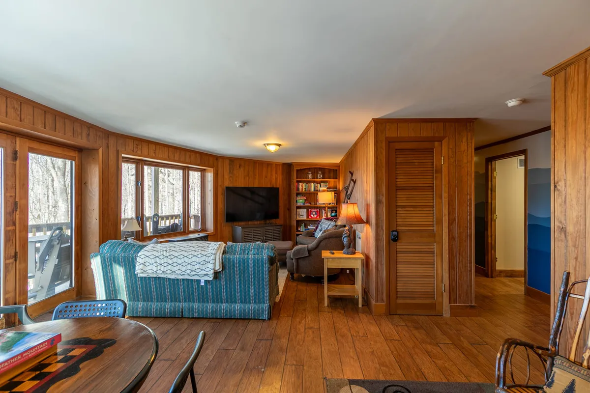 A cozy den featuring a blue striped couch, wooden shelves filled with books, large windows letting in natural light, a TV mounted on the wall, and a reading chair with a lamp. The furniture is arranged to encourage relaxation, socializing, or enjoying a movie night. Warm natural light streams in, creating an inviting atmosphere for unwinding after mountain adventures.