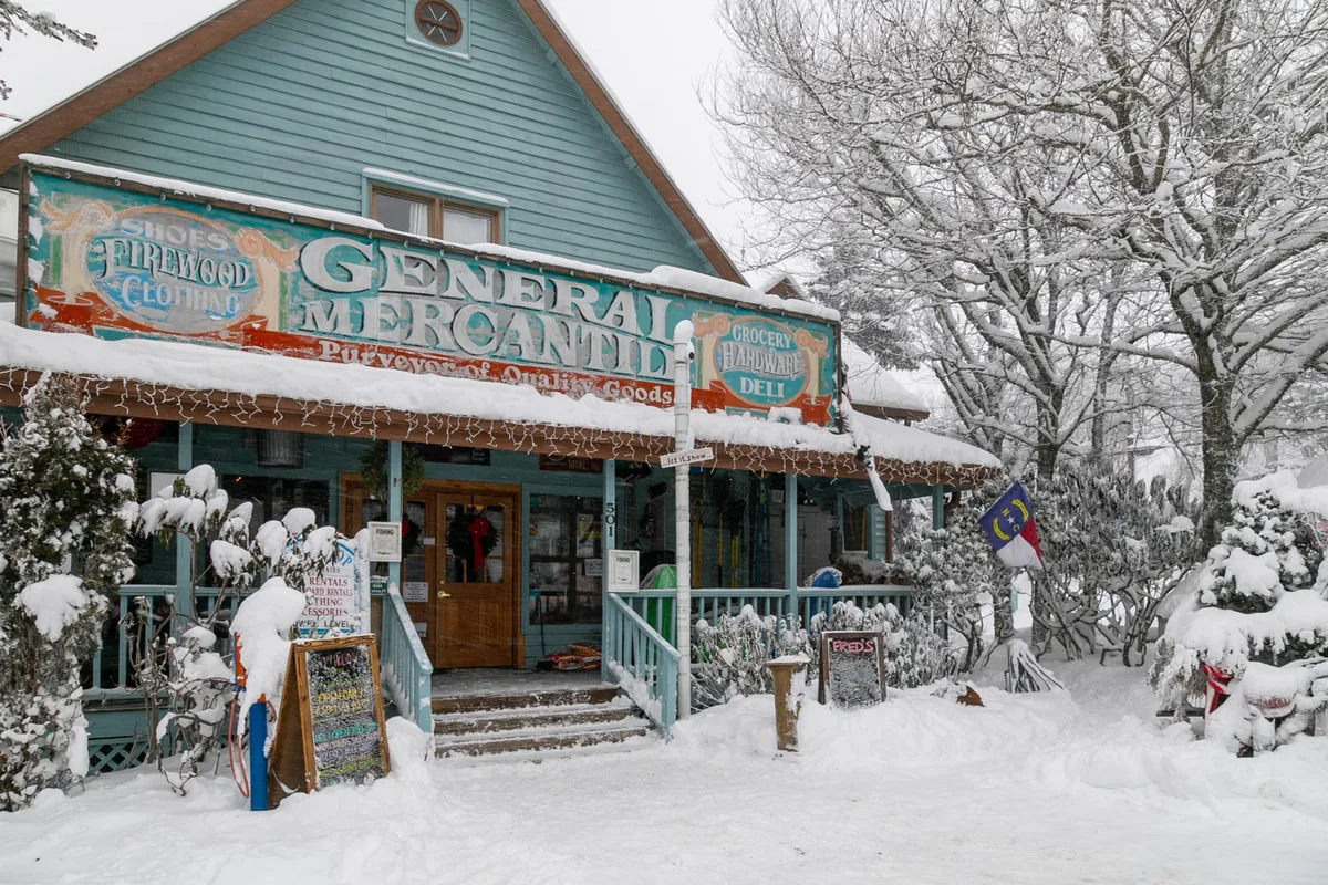 Fred’s General Mercantile’s snow-dusted porch, adorned with decorative lights and its classic sign, surrounded by trees. The welcoming atmosphere suggests a quaint, must-visit local spot for families.