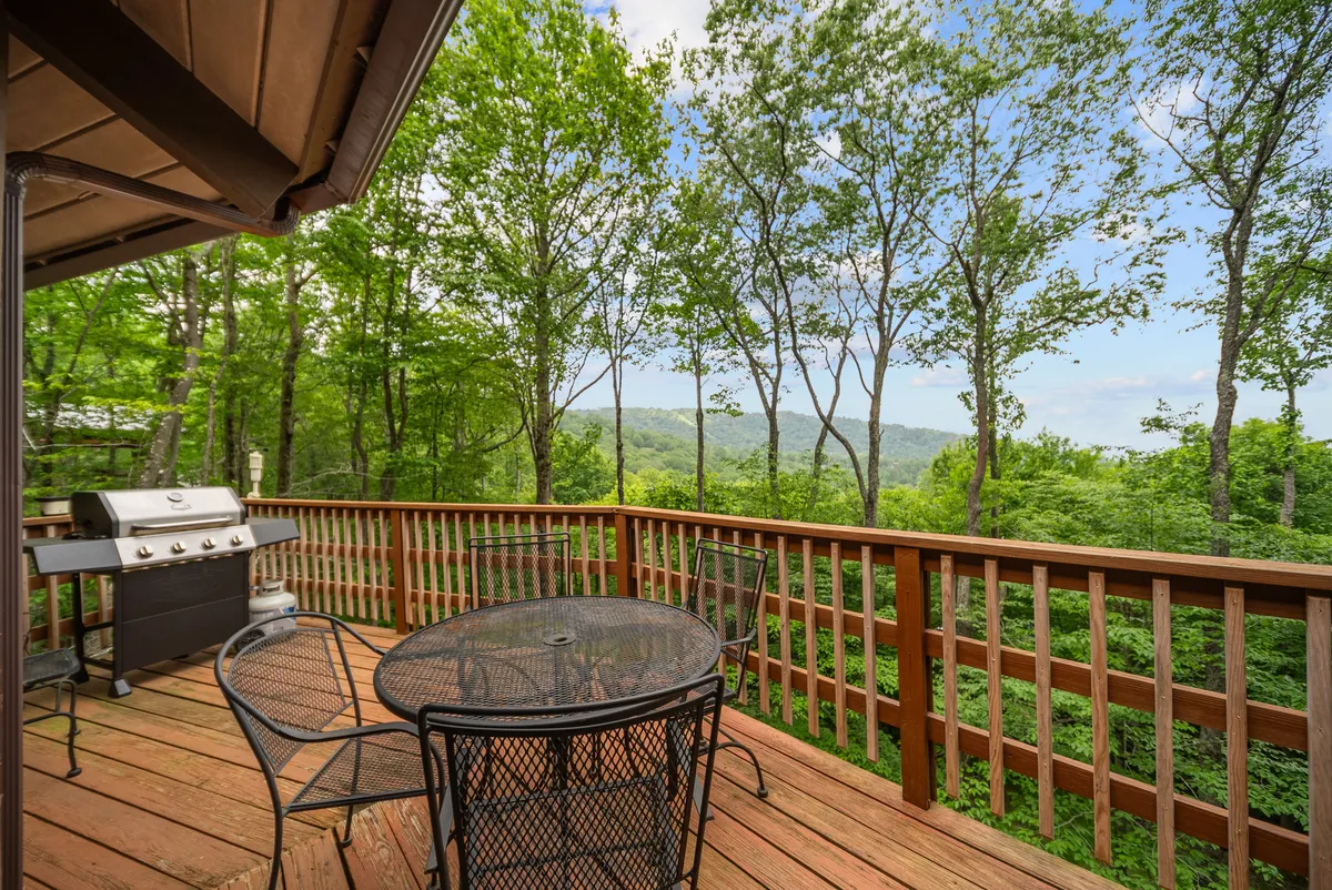 A spacious wooden deck outfitted with metal outdoor furniture and a grill, nestled amid lush greenery and overlooking distant hills. The setup suggests spaces for barbecuing, outdoor dining, and quiet relaxation, with panoramic views providing a scenic mountain backdrop.