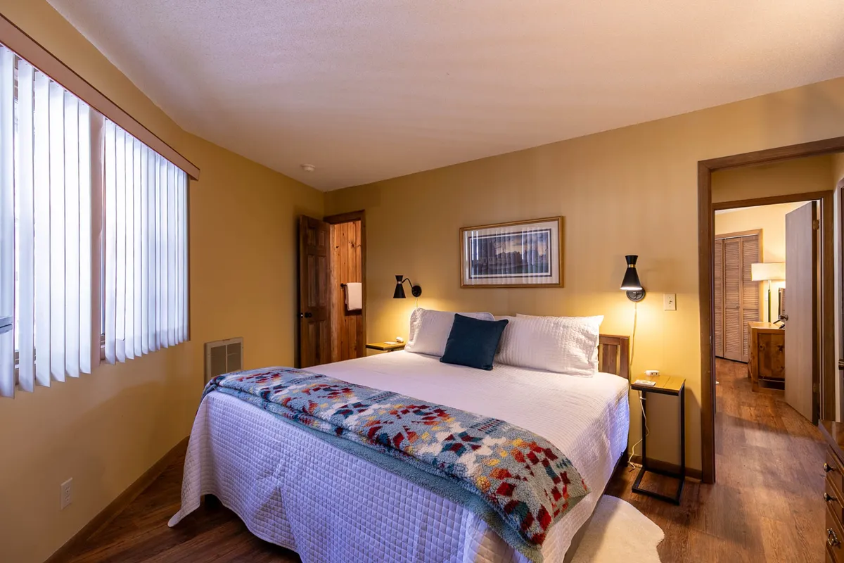 A comfortable bedroom decorated in soft tones, with a bed covered in a colorful blanket, wall art, and a nightstand with a lamp. The room feels bright and tranquil, emphasizing relaxation and quiet moments.