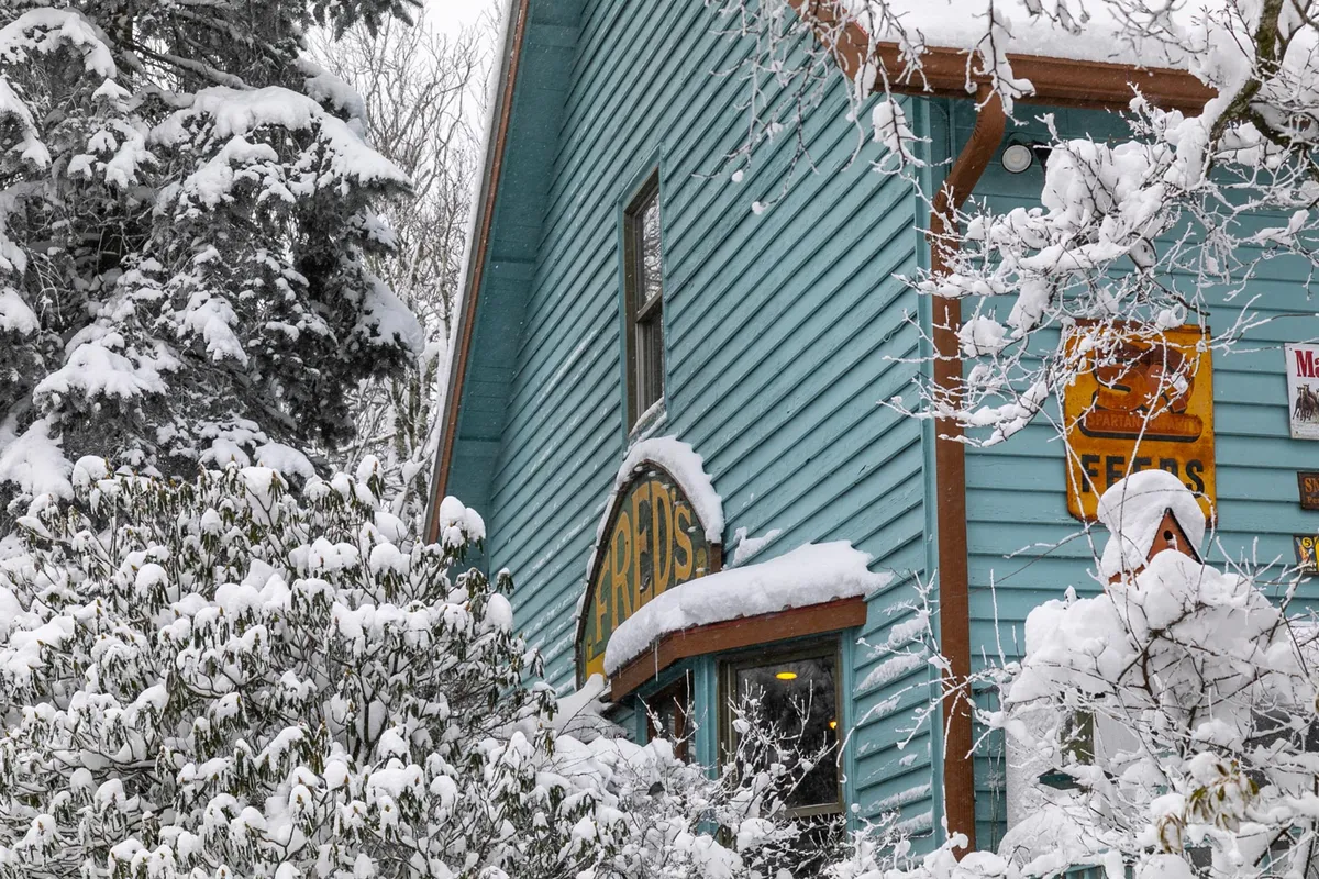 A cozy blue cabin with a snow-covered roof and decorative signage, nestled in a tranquil winter scene. The exterior suggests a welcoming, homey retreat, perfect for guests arriving with their pets after exploring the outdoors. The fresh snow and peaceful setting evoke the sense of a relaxing mountain getaway.