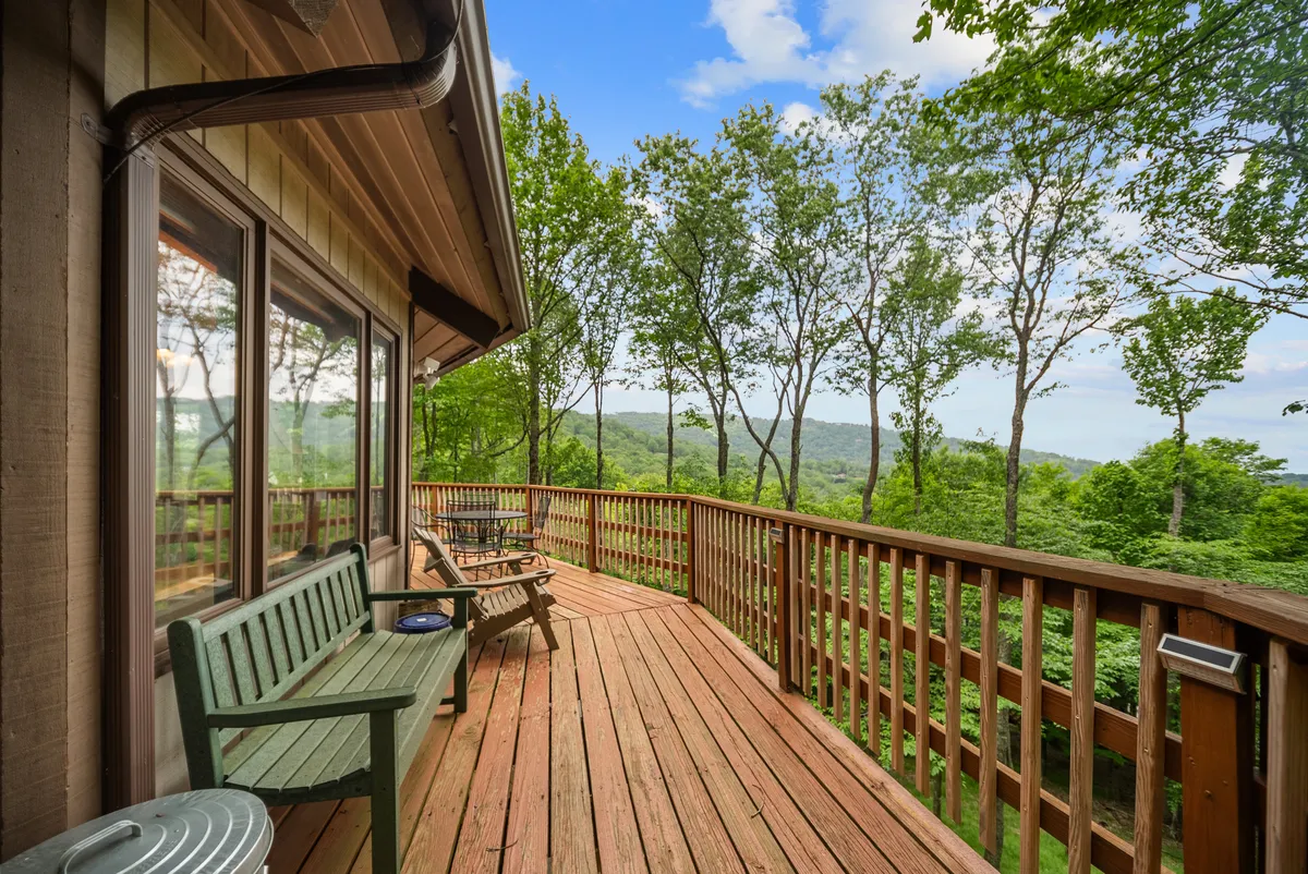 A spacious wooden deck with green benches and dining chairs, set against a lush backdrop of trees and sweeping mountain views. The space feels open and ready for family gatherings, wildlife watching, or simply soaking up the mountain air.