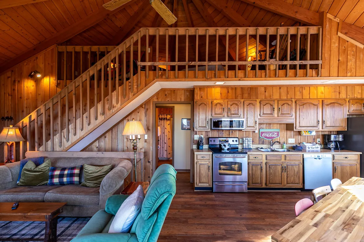 A view of the main living area with a prominent spiral staircase leading upward, kitchen space, living room seating, and warm wooden accents throughout. The staircase signals access to the loft above, hinting at the elevated retreat while tying the loft’s playful seclusion to the main home’s active energy.