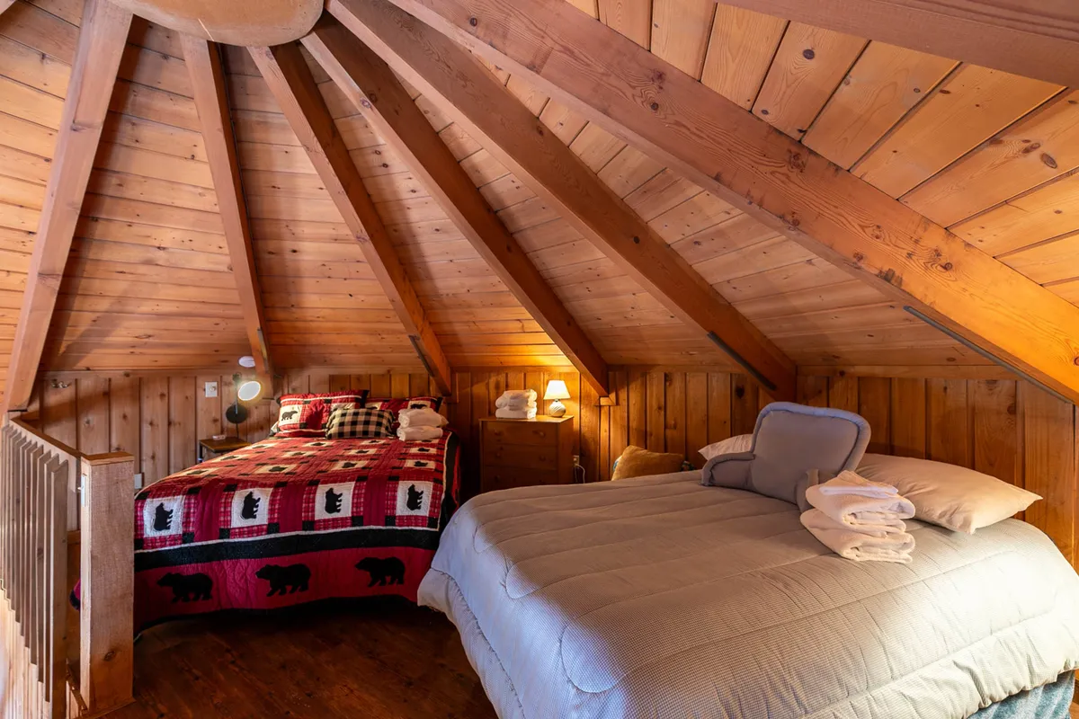 A cozy bedroom featuring wooden beams overhead, a bed with a plaid blanket, a second bed with a solid colored blanket, and a nightstand with a lamp. The wood accents and warm lighting create a peaceful, inviting atmosphere ideal for restful nights.